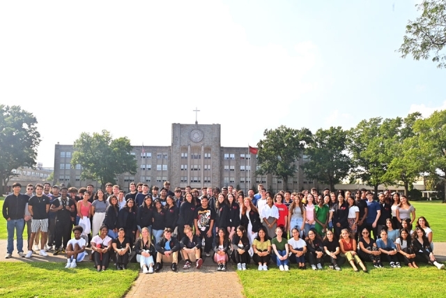 A large group of students poses outdoors on campus for a group photo, gathered in front of a university building on a sunny day.