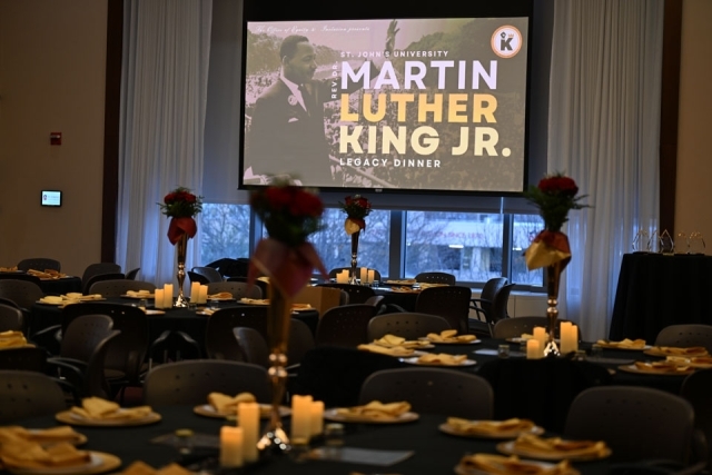 Decorated banquet tables set for the St. John’s University Martin Luther King Jr. Legacy Dinner, with candles, floral centerpieces, and a large event screen in the background.