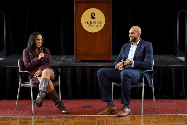 Two speakers seated on stage in conversation during a moderated discussion at St. John’s University.