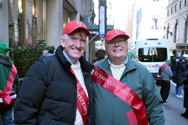 Two St. John’s University alumni wearing red sashes and hats smiling during a St. Patrick’s Day parade