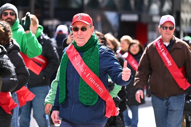 St. John’s University alumnus wearing a green scarf and red sash giving a thumbs up while walking in a St. Patrick’s Day parade