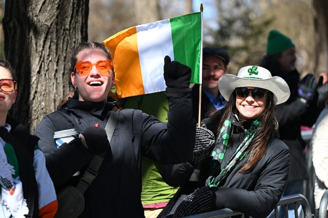 Spectators holding an Irish flag and smiling during a St. Patrick’s Day parade in New York City