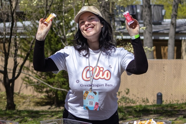 Student ambassador wearing a Clio Greek yogurt bars T-shirt smiles while holding up two sample bars at an outdoor campus event.