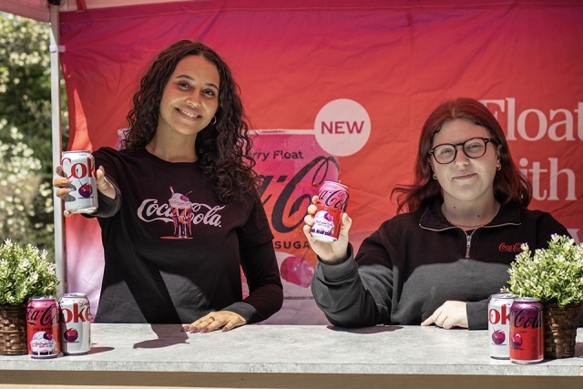 Two students stand at a Coca-Cola booth holding cherry-flavored soda cans in front of a bright promotional backdrop.