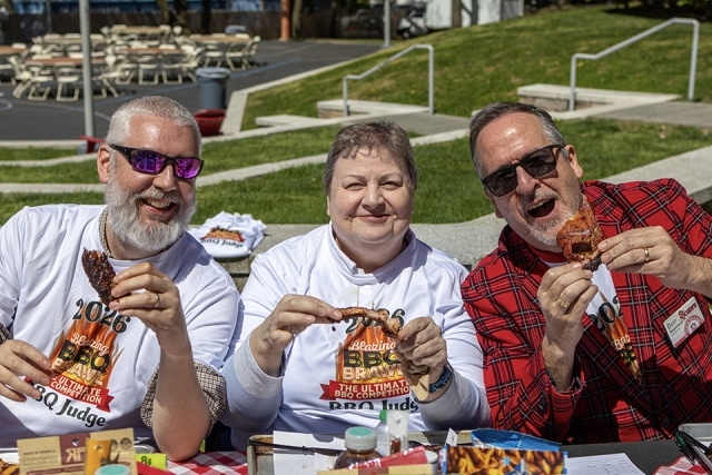 Three BBQ judges sit outdoors tasting ribs and smiling during a campus barbecue competition.