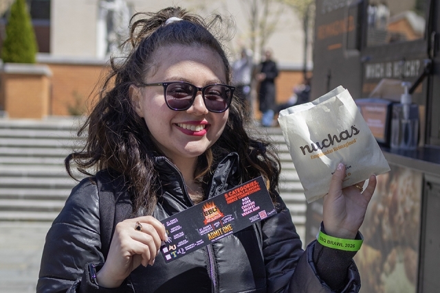 Student wearing sunglasses holds an event ticket and a bag of empanadas while standing on campus.