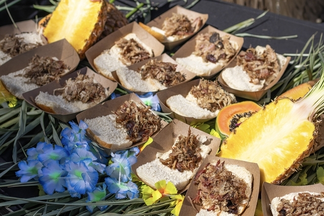 Display of pulled pork sliders arranged on a table with tropical fruit and decorative flowers.