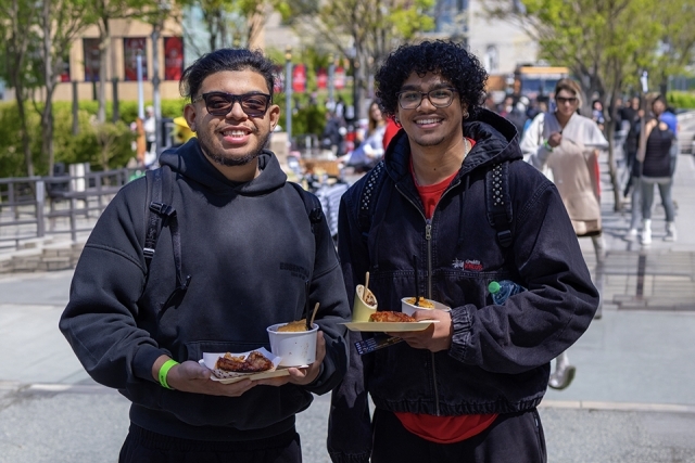 Two students smiling and holding plates of food while standing outdoors at a campus food event.