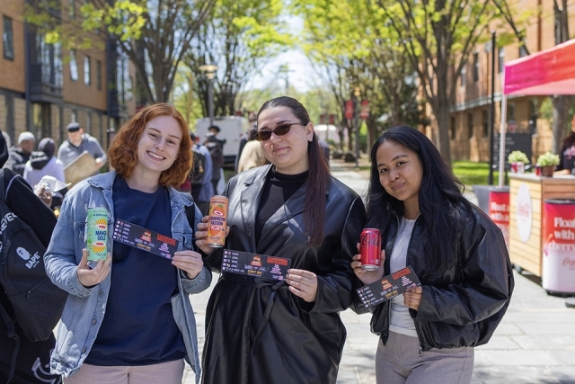 Three students pose together holding drinks and event tickets on a lively campus walkway.