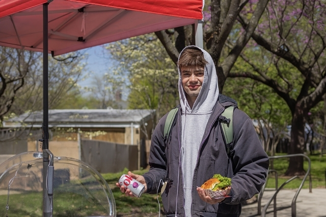 Student in a hoodie holds a soda can and a sandwich while standing under a tent at an outdoor campus food event.