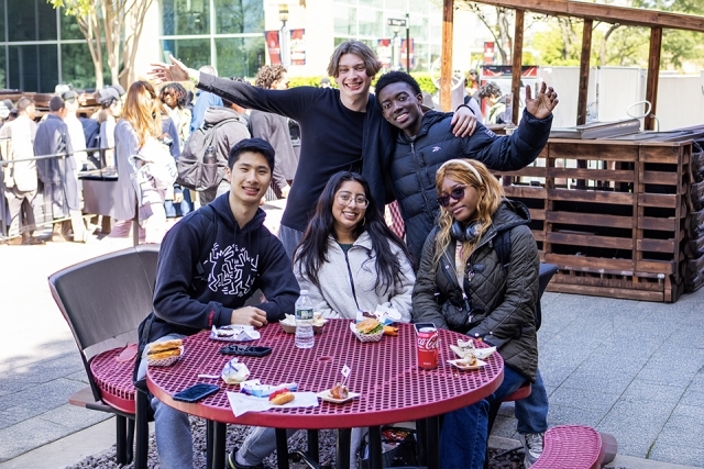 Group of five students gathered around a red outdoor table smiling and posing with food during a campus event.