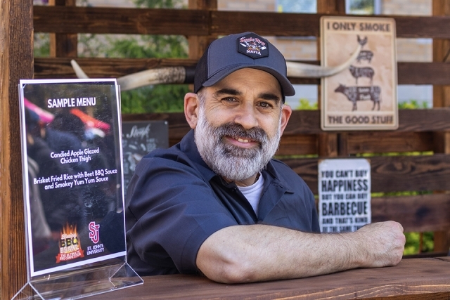 Vendor stands behind a wooden booth smiling, with a sample menu sign displayed at a campus BBQ event.
