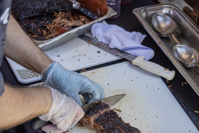 Close-up of a gloved hand slicing smoked brisket on a cutting board at a barbecue station.