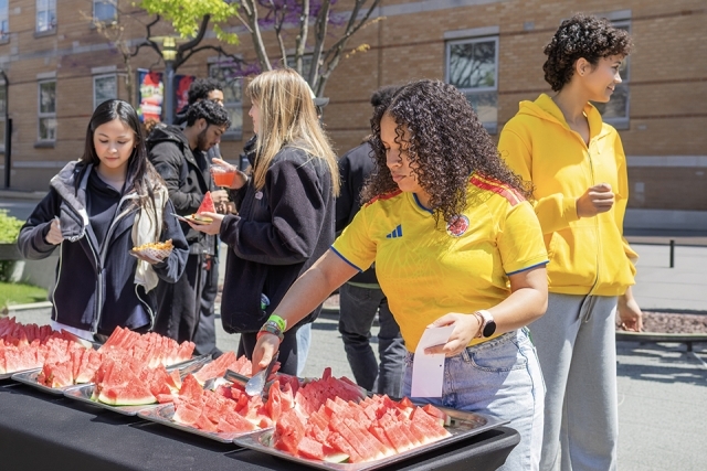 Students gather around a table selecting fresh watermelon slices at an outdoor campus event.