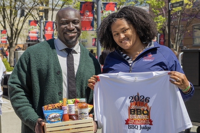 Two staff members smile while holding a prize basket and a “BBQ Judge” shirt at a campus event.
