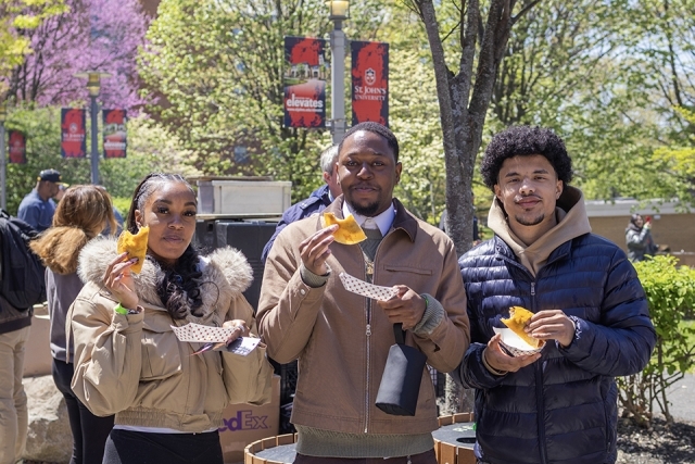 Three students stand outdoors holding empanadas and smiling during a campus food festival.