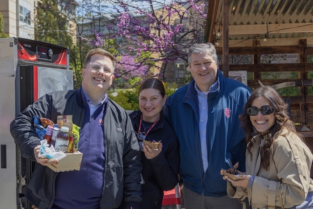Group of four staff members smiling and holding food samples while standing near a campus food truck.