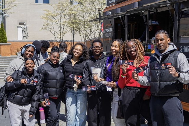 Large group of students pose together holding event tickets in front of a campus food truck.
