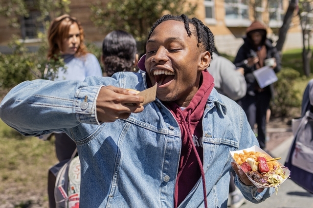 Student takes a bite of a food sample while holding a tray of loaded fries at an outdoor campus event.
