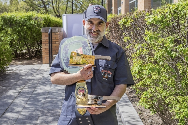 Barbecue competitor smiling while holding multiple awards, including a championship belt and cleaver-shaped trophies, outdoors on campus.