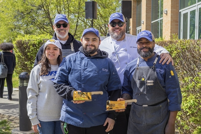 Group of chefs and team members posing together and holding BBQ competition trophies at an outdoor campus event.