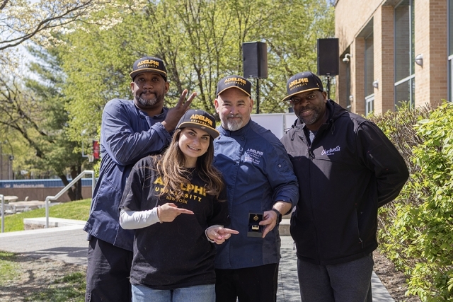 Team wearing Adelphi University apparel poses together, smiling and holding an award during a campus BBQ competition.