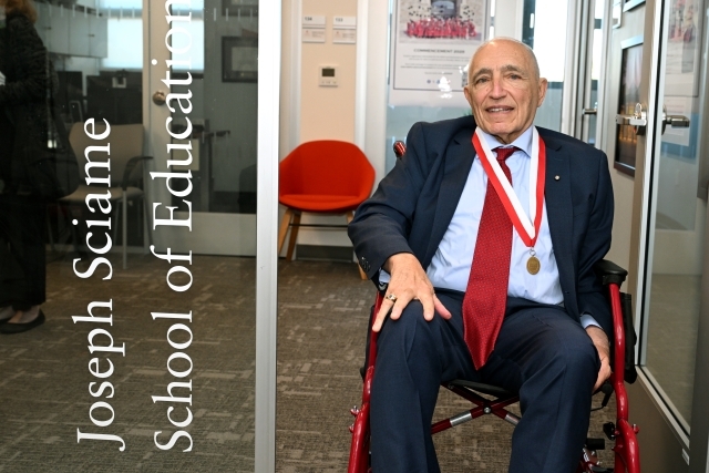 Joe Sciame seated outside door labled Joseph Sciame School of Education