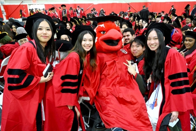 Graduates in red commencement gowns pose with the St. John’s University mascot during a graduation ceremony inside a crowded arena.