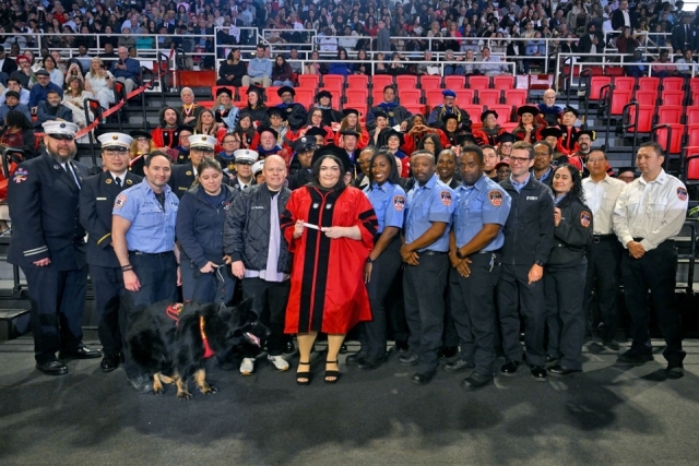 Graduate in academic regalia stands with emergency service professionals and a service dog during a St. John’s University commencement ceremony.