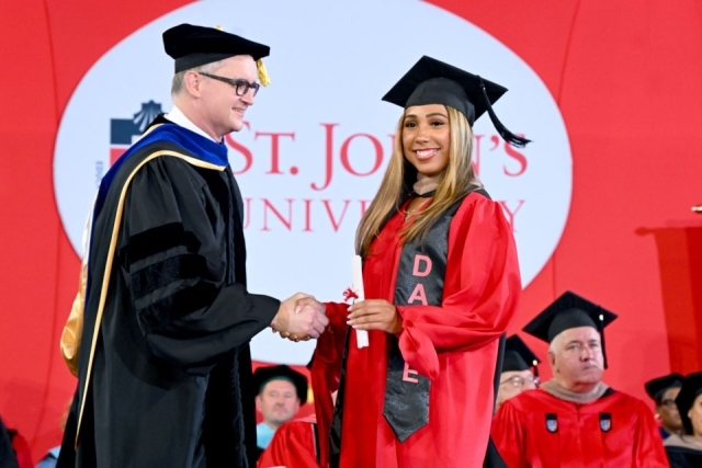 Graduate wearing a decorated stole smiles while receiving a diploma during St. John’s University commencement.