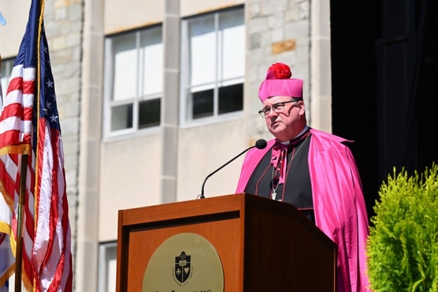 University leader delivers a commencement speech at a podium during an outdoor St. John’s University graduation ceremony.