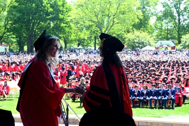 Two graduates shake hands on stage during St. John’s University commencement ceremony with graduates seated in the background.