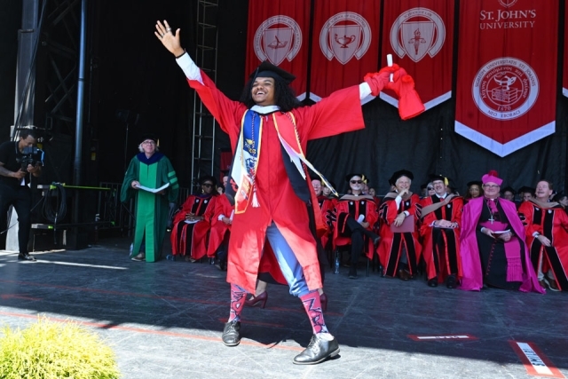 Graduate celebrates on stage with arms raised during St. John’s University commencement ceremony.