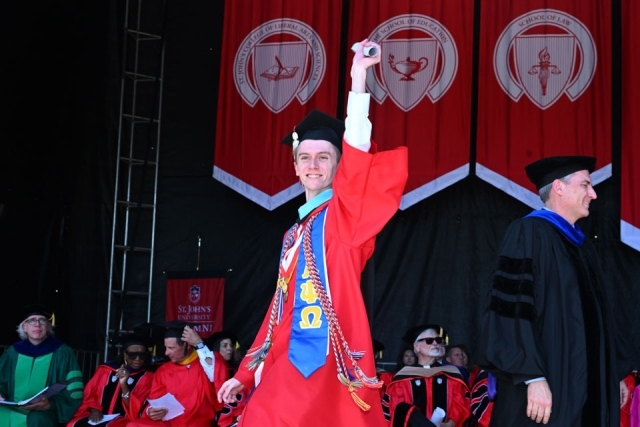 Graduate celebrates on stage with arms raised during St. John’s University commencement ceremony.