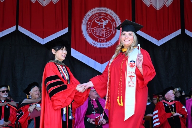 Nursing graduate poses with diploma while shaking hands with faculty member during St. John’s University commencement.