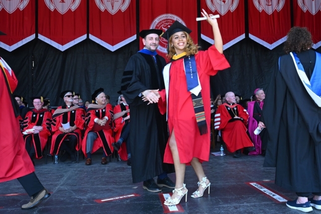 Graduate waves diploma while celebrating on stage during St. John’s University commencement ceremony.