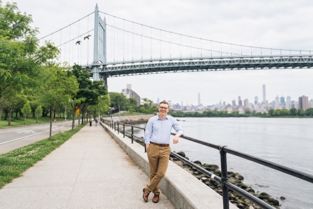 Hunter Igoe stands on a tree -lined riverside promenade in new York City with a bridge and the city's skyline in the background.