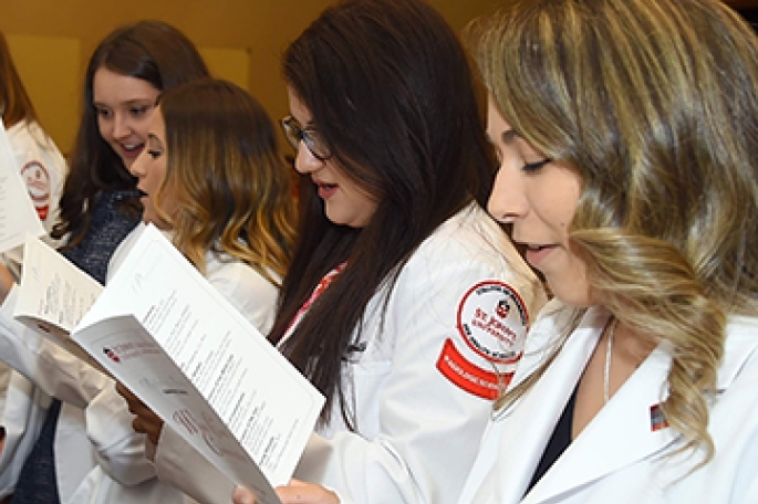 Four female St. John’s University students wearing white lab coats look at the event program for the pharmacy white coat ceremony.