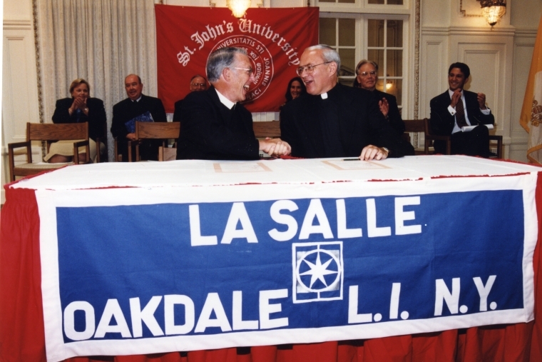 Brother A. Jerome Corrigan, left, shakes hands with Rev. Donald J. Harrington, CM, right, sitting at a table draped with a La Salle banner, and a St. John's University banner on the wall