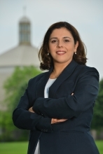 Professional head shot of Maha Saad. She is standing in front of St. Thomas Moore church on a cloudy day. Her arms are crossed in front of her. She is wearing a navy blue blazer, light blue blouse. She is smiling.