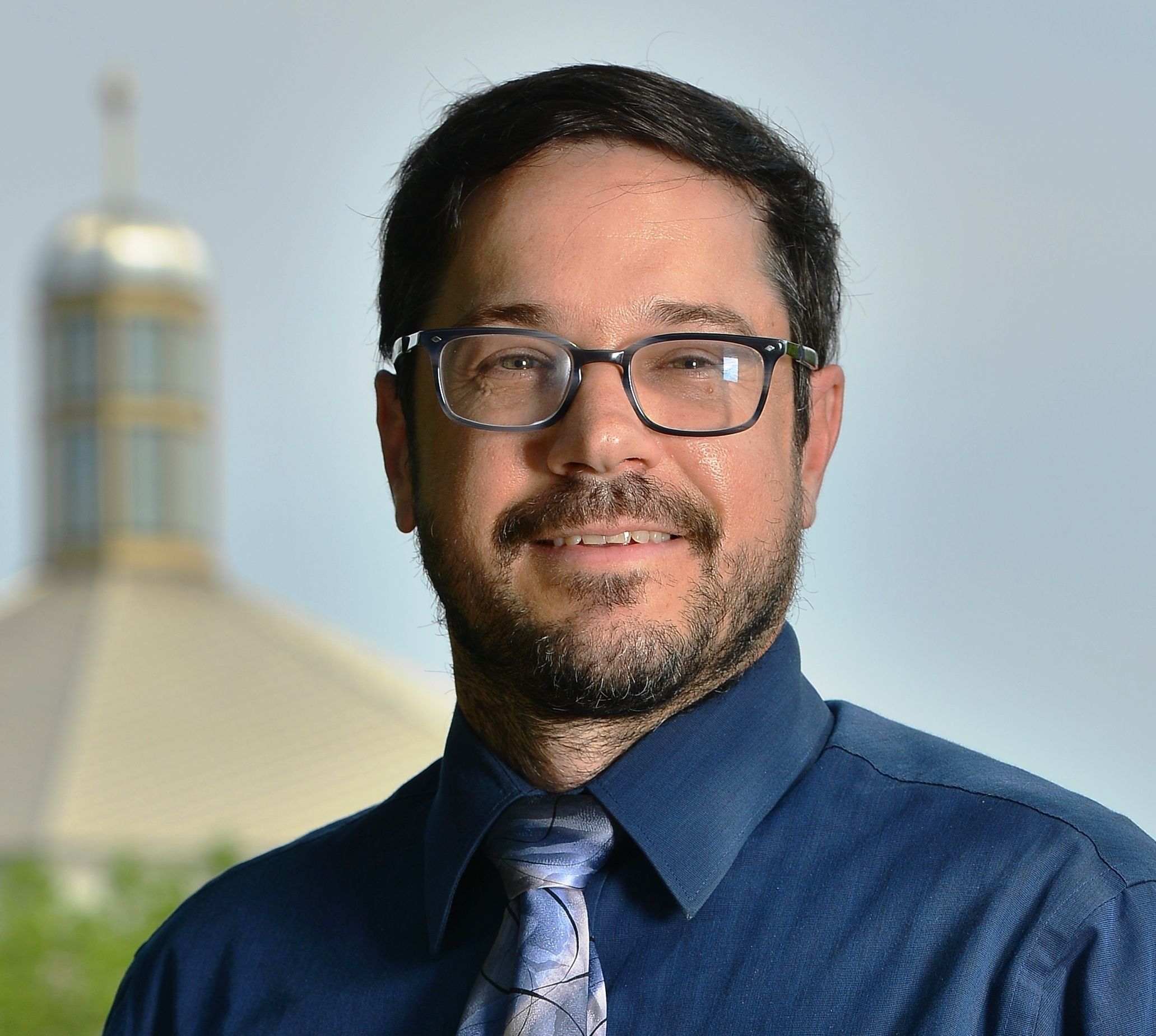 Head shot of Dr. Martino. He is wearing glasses, a dark blue shirt and blue tie, with St. Thomas Moore church in the background.